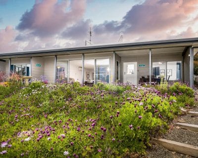Elegant front view of Amani House at Emu Bay, Kangaroo Island, designed to accommodate 2-9 guests. This beautifully renovated home boasts expansive sliding doors that frame stunning vistas from its elevated position. The property is equipped with a modern kitchen and a new, fully accessible second bathroom, making it ideal for a relaxing getaway.